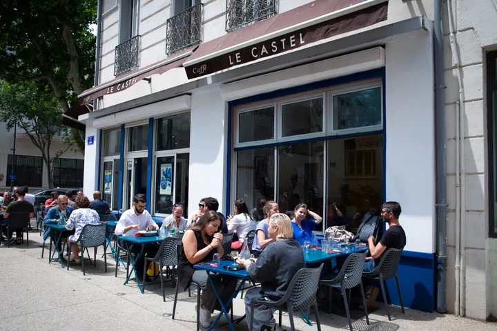 La façade blanche de la Brasserie Le Castel et sa terrasse au soleil, clients attablés aux tables bleues